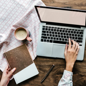 Photo of a person sitting at a table using a laptop with a notebook and cup of coffee