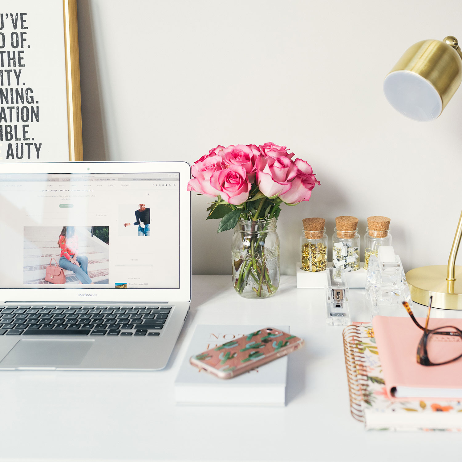 Vibrant photo of a desk filled with a laptop, vase of flowers, notebook, lamp, and glasses